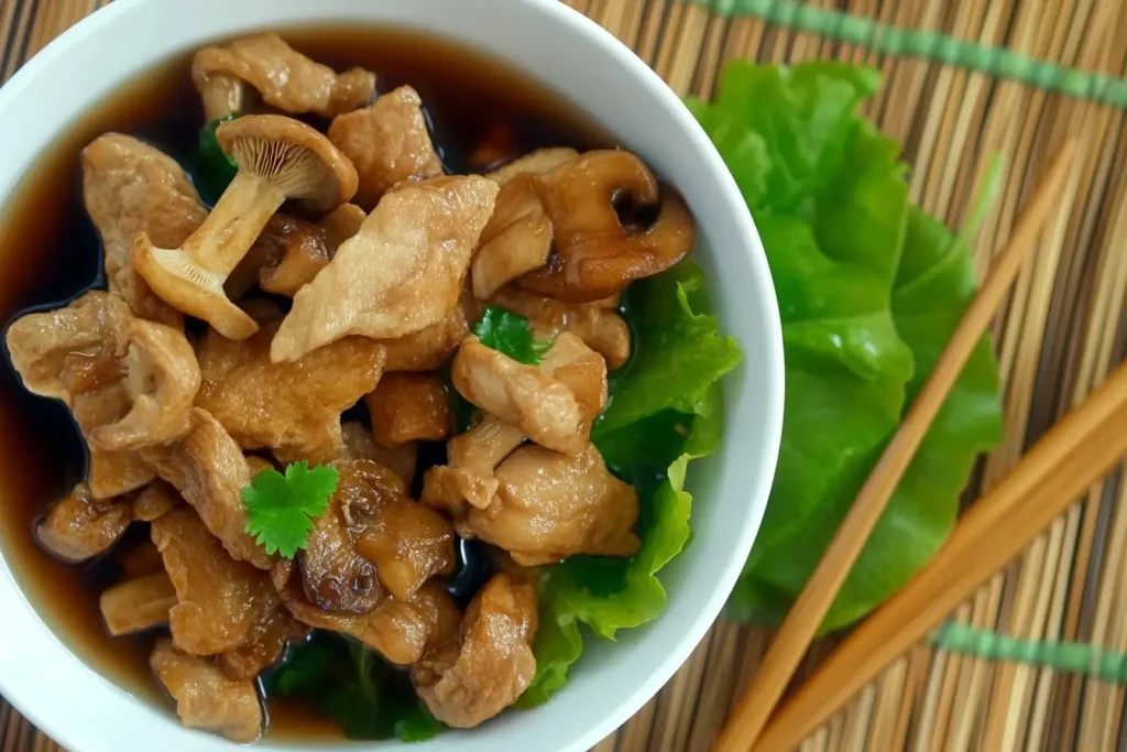 A bowl of chicken and mushroom stir-fry, served with fresh lettuce and a flavorful soy-based sauce, garnished with coriander, on a bamboo placemat with chopsticks on the side.

