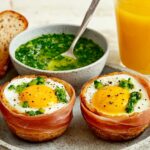 Three prosciutto-wrapped egg cups filled with baked eggs and topped with a vibrant green herb sauce, accompanied by bread slices and a bowl of herb sauce.